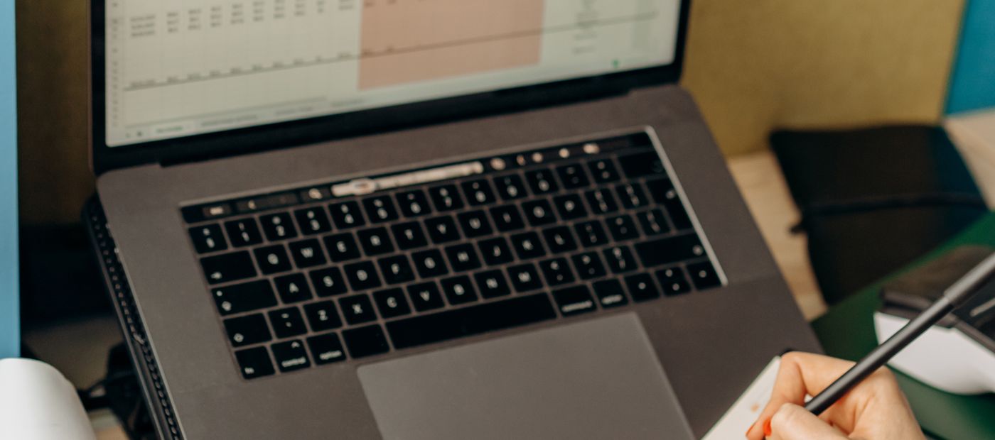 A spreadsheet open on a laptop, a hand with a pen checking a row against paper