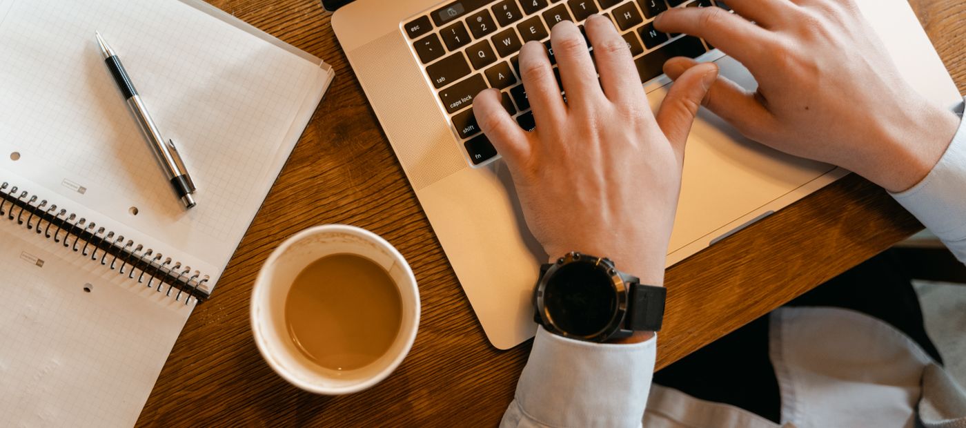 Hands typing on a laptop beside a coffee and an open notebook